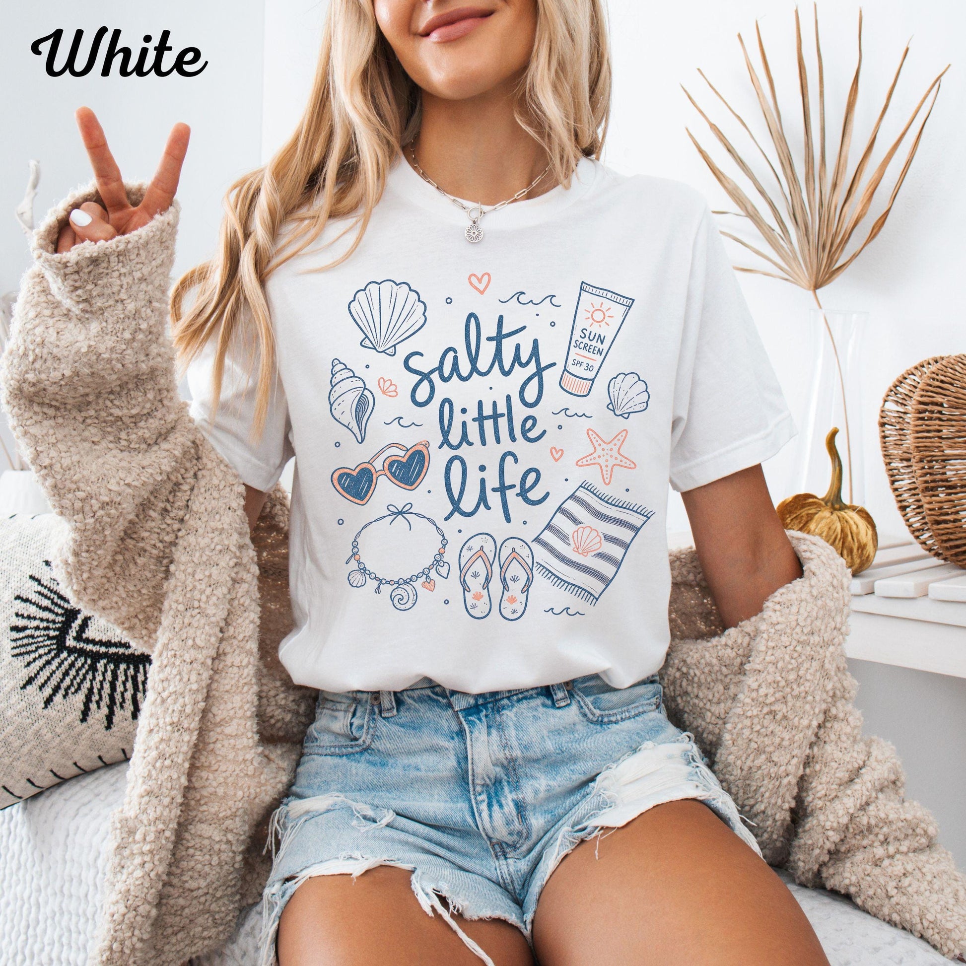 May include: White t-shirt with the text "salty little life" and beach-themed illustrations. The design includes seashells, sunglasses, a necklace, flip-flops, sunscreen, and a striped towel. The word "White" is in the top left corner.